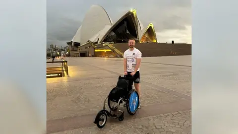 Billy Whiston is standing with his wheelchair in front of the Sydney Opera House, at the start of his journey.