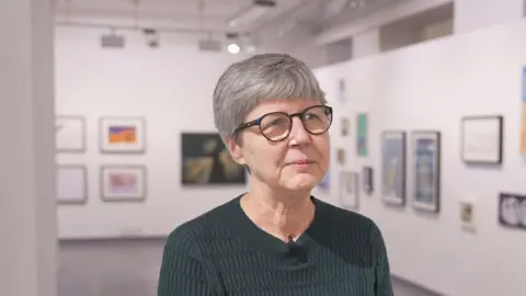 Elisabeth Gibson, has short, grey hair and is wearing a dark top. She is sitting down, wearing glasses and is in an art gallery or studio, with paintings and art hanging on the walls around her. 
