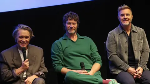 Getty Images Mark Owen, Howard Donald and Gary Barlow are seated and holding microphones. Owen, who sits to the left, is smiling and holding his microphone up to his mouth. 