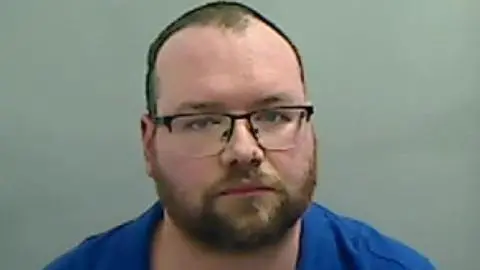 Andrew Barber, who wears black glasses and has a short beard, looks up at the camera. He wears a blue shirt and has no expression. His short dark hair has receded to the top of his head.