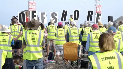 The protesters were holding placards that read 'Our communities are being killed'. He is wearing a high-vis jacket "offshore4sure" written on the back