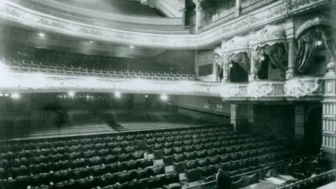 Manx National Heritage A black and white image of the view from the stage of the theatre, rows of wooden seats make up the stalls, and more can be seen in the circle too. There are individual boxes close to the stage on the circle-level. Ornate designs can be seen on the outside, which connect to front of the balconies.