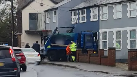 A photo of the collision in Canvey. It shows a blue skip lorry resting in the front of a house. A police officer can be seen stood next to the lorry speaking with another person in an orange hi-vis. Other people can be seen behind the lorry. Cars are driving past while a police car can be seen parked nearby.