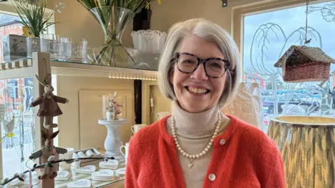 Jenny Kirk/BBC Suzi is standing in her shop.
She has a bob like length hair and is wearing glasses.
She is also wearing a mid orange coloured cardigan and a beige sweater underneath.
In the background, her shop artefacts can also be seen