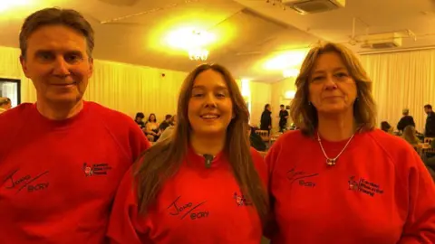 A man and two women looking at the camera. They are wearing bright red polo jumpers with the logo "Cardiac Risk in the Young" printed on them. The words "Jono @ CRY" are also printed on them. They are standing in a white marquee with about a dozen people sitting or stands at tables in the background.