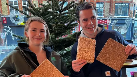 BBC Caelo and Hugo stand indoors in front of a Christmas tree, each holding rectangular boards made from compressed Christmas tree material. Both are smiling at the camera.