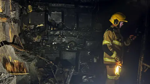 Shropshire Fire and Rescue Service A firefighter with a yellow helmet is in the charred property, carrying a torch and holding equipment.