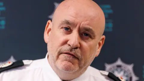 PA Media A man with a bald head and grey beard wearing a white shift with black shoulder pads bearing the crest of the Police Service of Northern Ireland. 