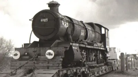 Buckinghamshire Railway Centre A black and white photo of the train when it was rescued from a scrapyard, it is covered in graffiti ad sat on the back of a flatbed lorry