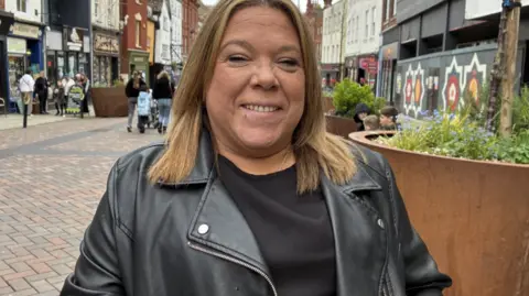 A lady with light brown hair and a piercing below her bottom lip. She's wearing a black leather jacket with a black top underneath. She's stood on a high street with shops and people walking behind her.
