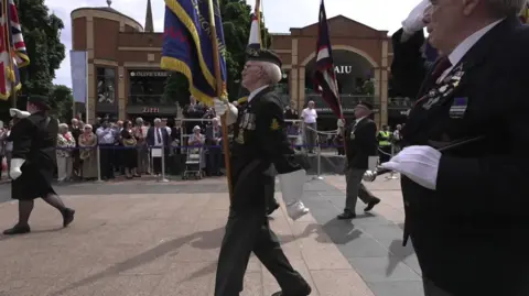 BBC A woman and several men in black suits and wearing white gloves hold flags as they walk along the pavement. In the background, a crowd stands in front of a brown-brick building.