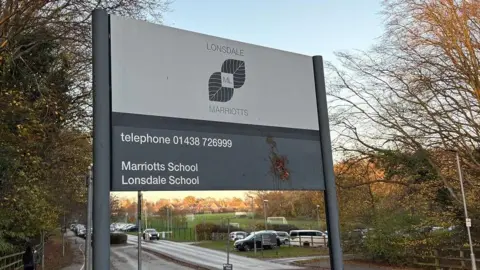 Martin Heath/BBC A sign at the entrance to school with "Lonsdale School" in grey lettering with a grey leaf logo. There is also a telephone number in white lettering on a grey background. There are trees on both sides of the sign and a car park beyond it.