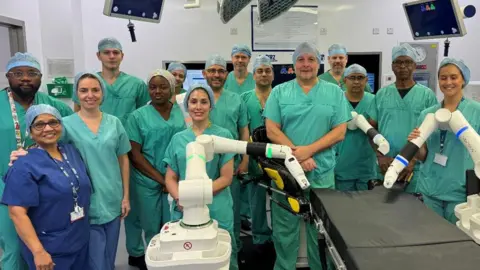 A dozen men and women wearing turquoise scrubs and blue hair nets stand around the new Versius surgical robot. The three white robotic arms are positioned over a black bed where the patient would lay. 