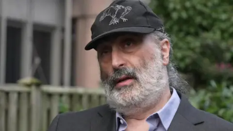 Mark Nygate, a man with a grey beard, wearing a dark suit and a baseball cap.