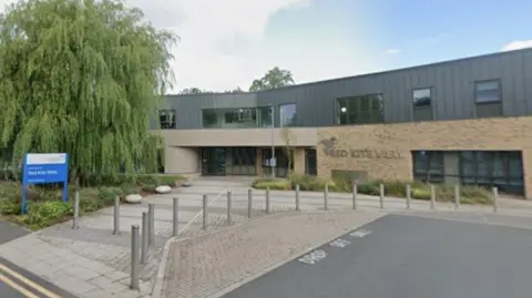 Google The outside of a modern build NHS building. Bollards and a path leading up to the front door, as well as a sign indicating the name of the premises can be seen.
