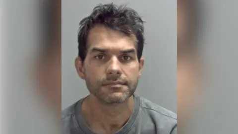 A police custody photo of Tancredo Bankhardt looking directly at the camera. He has short dark hair and stubble, and is wearing a grey T-shirt.