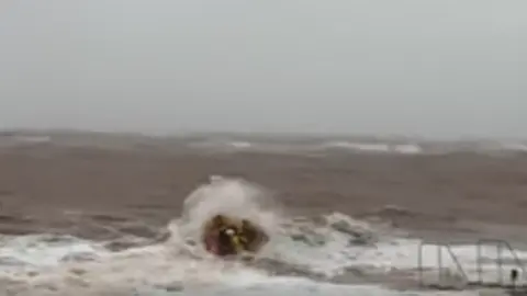 Image shows lifeboat crew heading out on a windy day to rescue two wingsurfers. The volunteer crew, dressed in yellow, are on a small orange boat. They are heading out into stormy seas. Large waves are crashing over the boat carrying the crew.