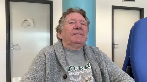 John Fairhall/BBC David Bancroft, 76, is sitting down wearing a grey t-shirt and cardigan while waiting to see spinal specialists in Ipswich Hospital