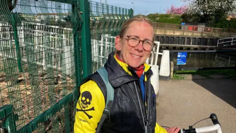 Claudia has brown hair and glasses on. She is wearing a black and yellow jacket and has a bicycle with her. 