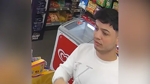 A young man with dark hair in a white Calvin Klein top stands at the till in a corner shop.