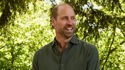 Prince William is wearing a dark green shirt. He is looking away from the camera and smiling. The background is out of focus but has greenery and trees in the background. 