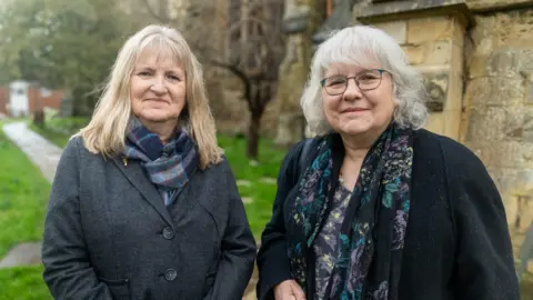 Ian Palmer/BBC Two woman look in to the camera with slight smiles. The woman on the left has long blonde hair. She wear a blue tartan scarf and a blue grey wool coat. The woman on the right has short grey hair with a wave. She wears green glasses. She has a black coat on and is wearing a floral top with a floral scarf in black, green, yellow and purple.