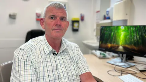Simon West is sat in an office, he is wearing a patterned shirt and is in front of a computer. 