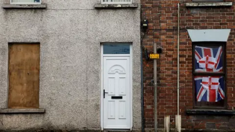 A faded white walled townhouse is attached to an old red bricked home. The faded white house has a white PVC door and its downstairs window has been boarded up. The red bricked house has a red, white and blue Union Jack flag in the window with two smaller Ulster Banner flags.