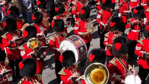 An aerial shot of the Coldstream Guards marching through Berwick. They are wearing full uniform with bearskin hats and bright red military jackets. All are playing musical instruments including wind instruments and drums.