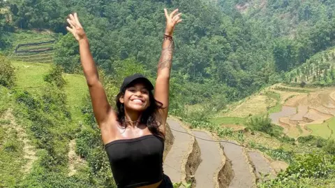 Family handout Juliana Marins raising both hands in the air, in a black cap and black tube top. In the background are green paddy fields.