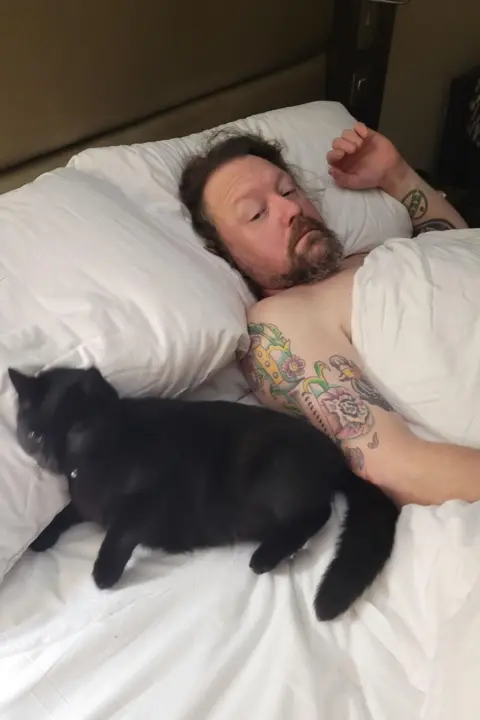 Sarah Shepherd A man laying in bed looking at the black cat laid next to him