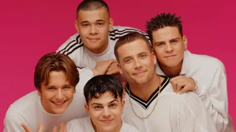 Getty Images All five band members smile posed in a huddle together infront of a bright pink backdrop. They all have white t shirts on. 