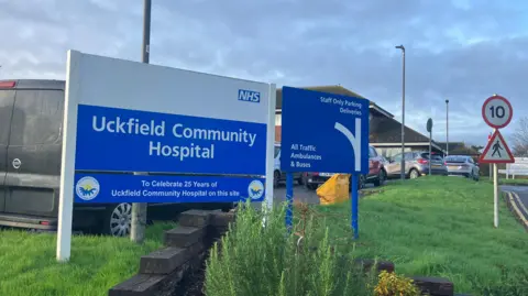 A blue sign at the entrance to Uckfield Community Hospital, welcoming visitors to the site. The hospital building can be seen in the background.