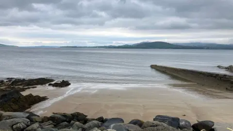 A sandy slope down into the blue sea. There are grey and black rocks on the beach and a stone wall along the side. There are hills in the background.