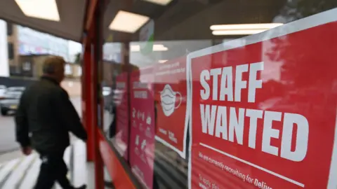 An image of a man walking past a red sign in a window saying staff wanted