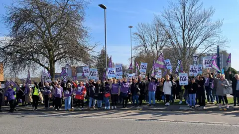 Lots of people with flags saying "Pay Fair". They are standing outside on a sunny day. 