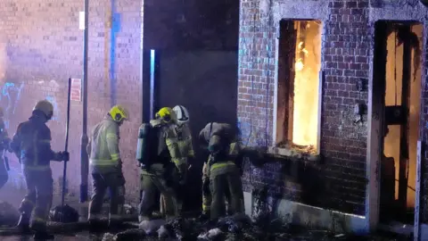 PA Media Firefighters work outside a building damaged by fire, with flames visible through a window and doorway. The exterior walls are blackened by smoke as emergency crews respond at the scene during the night.