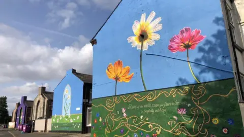 Liverpool City Council Murals on the end of rows of terraced houses feature grass, blue sky and various flowers in different colours including orange, pink and blue.