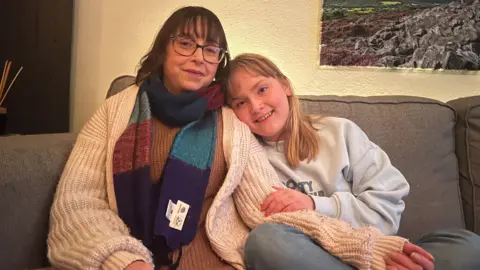 Jen left a woman with brown hair and glasses, sat on a sofa wearing a cardigan jumper and scarf next to her daughter Emberly Heppenstall, a young girl with blond hair and a white sweatshirt, resting her head on her mother's shoulder.