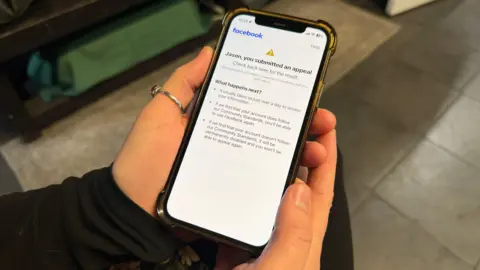Two hands holding a smart phone. The phone shows a warning message which says 'Jason you have submitted an appeal'. The Facebook logo is in the top left hand corner of the screen.