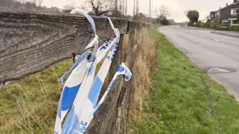 Ragged police tape sitting on top of a wooden fence. A grass verge and suburban road sit to the side of the fence.
