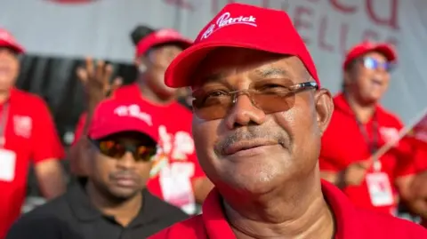 Reuters A head and shoulders shot of Patrick Herminie. he is smiling and wearing a red campaign baseball cap. Supporters can be seen behind him - they are out of focus.