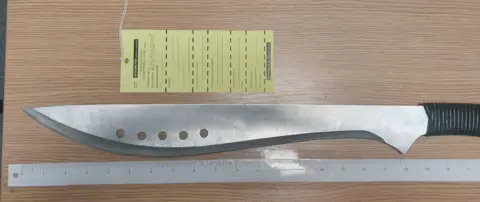 Derbyshire Police A silver machete with black handle on a wooden surface with a ruler next to it