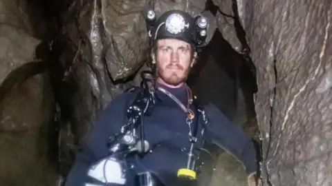 George Linnane inside a cave and looking at the camera. He is wearing a wetsuit and a safety helmet.