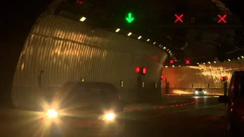 BBC Several cars with headlights on drive through a dark, curved road tunnel with illuminated green arrows and red crosses above their heads.