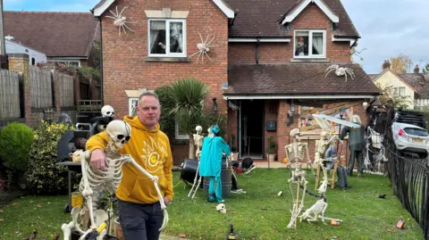 Anthony Jackson in a yellow hoodie stood in his front garden surrounded by skeletons and other Halloween decorations. 