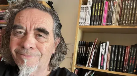 Richard Jones Richard has short curly hair and a beard and is looking at the camera on the left smiling. There is a bookshelf behind him of books.