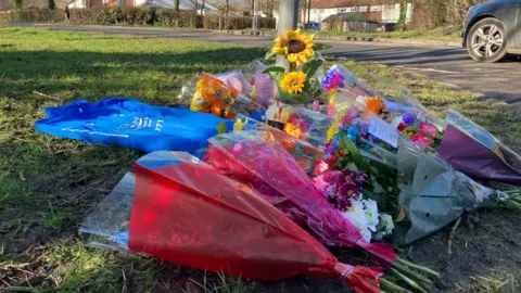 Sam Read/BBC Bunches of flowers are lying on grass at the side of the road. Some have messages written on them