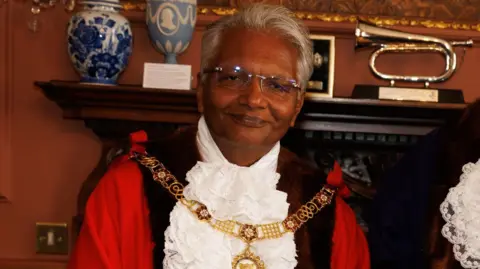 Dr Bharat Pankhania. He has short grey hair and glasses and is wearing civic regalia. He is looking at the camera and smiling.