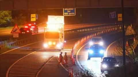 Night-time road repairs with men in hi vis in foreground and lots of cones and an illuminated lorry